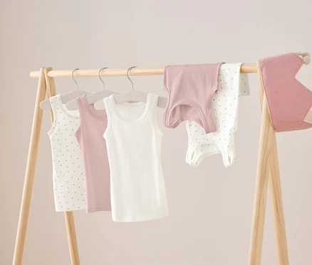 Auf einem Holzständer hängen drei Unterhemden in Weiß, Rosa und Weiß mit Punkten, sowie ein rosa Hemd und ein weißer Body mit Punkten. Rechts hängt eine rosa Krone.