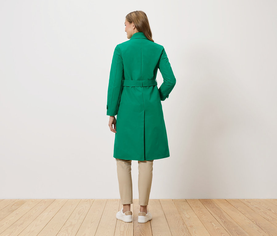 Eine Frau steht mit dem Rücken zur Kamera und trägt einen grünen Mantel mit Gürtel, beigefarbene Hosen und weiße Turnschuhe.