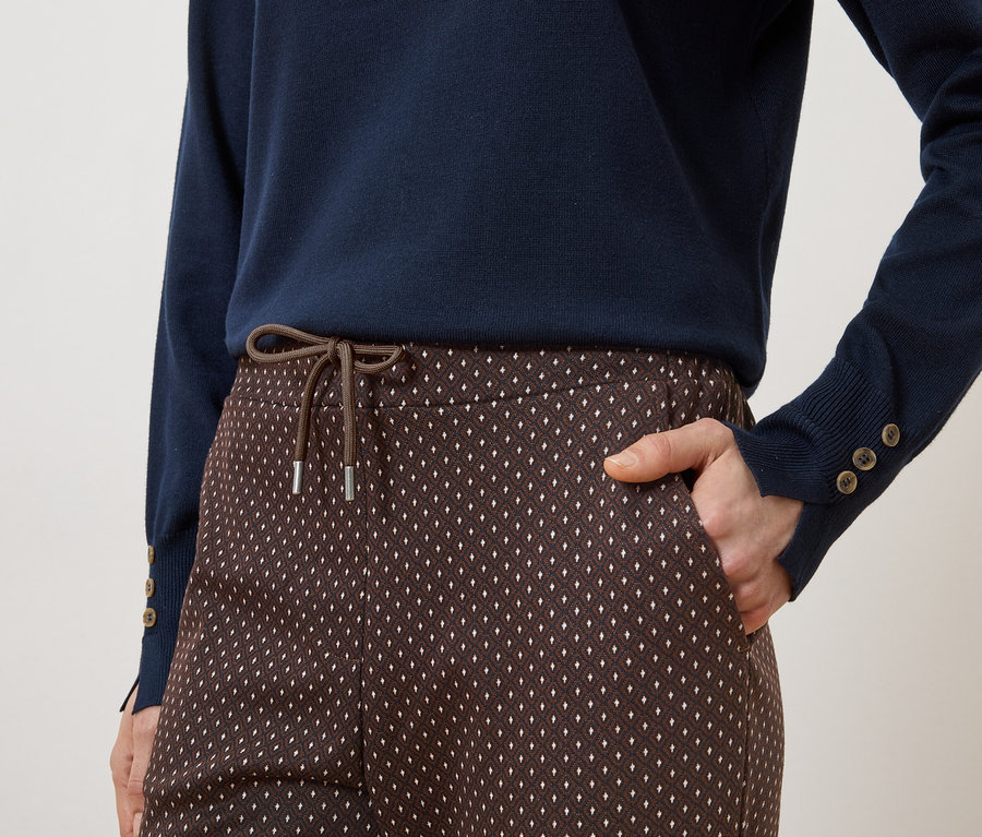 Person mit marineblauem Pullover und braun gemusterter Hose mit Kordelzug und Hand in der Hosentasche.
