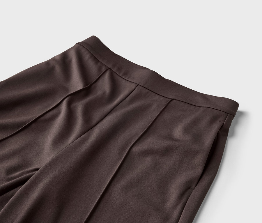 Nahaufnahme von der Taille einer dunkelbraunen Hose mit Bundfalten und einer Seitentasche vor einem hellgrauen Hintergrund.
