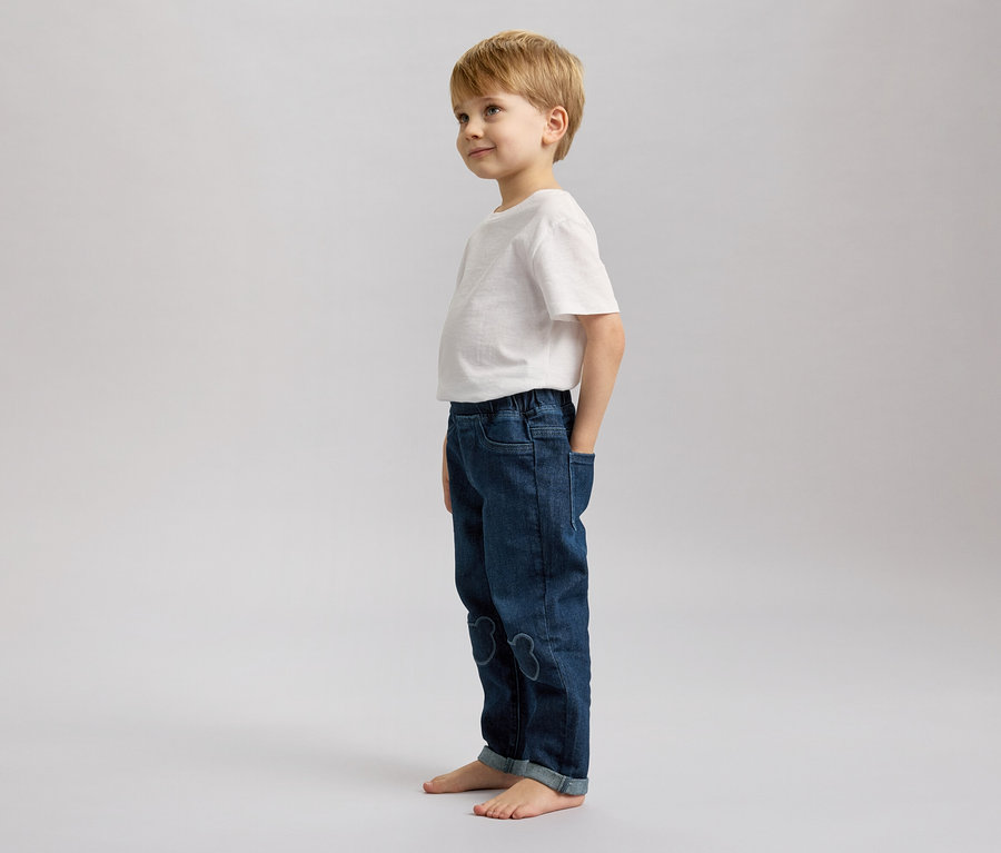 Ein Junge mit blonden Haaren steht barfuß in Jeans und einem weißen T-Shirt da und schaut nach links.