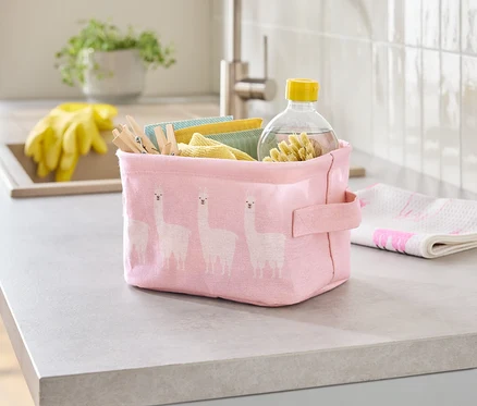 Auf einer Küchentheke steht eine rosa Stoffbox mit Lama-Muster, gefüllt mit Reinigungstüchern, Wäscheklammern und einer Flasche Reiniger.