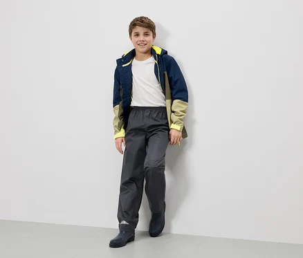 Ein lächelnder Junge in Regenjacke, Hose und Stiefeln lehnt an einer Wand.