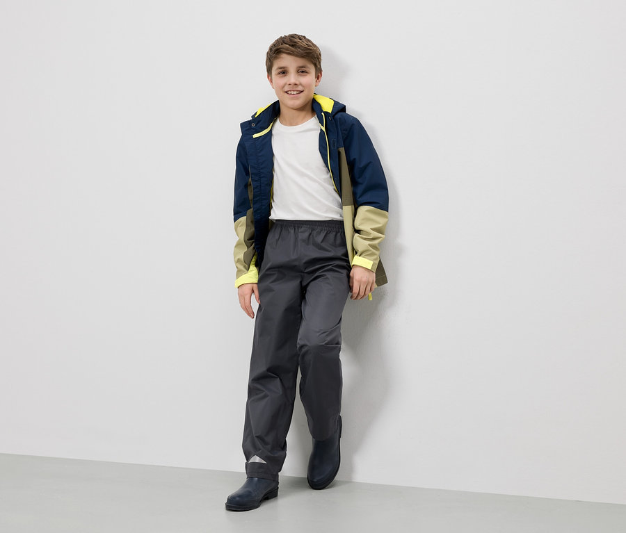 Ein lächelnder Junge in Regenjacke, Hose und Stiefeln lehnt an einer Wand.