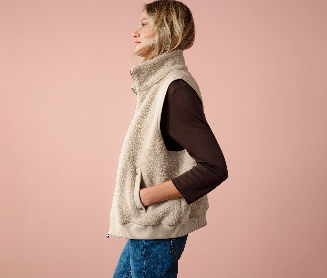 Frau mit Street One Teddyweste, braunem Oberteil und Jeans, seitliche Ansicht vor rosafarbenem Hintergrund.