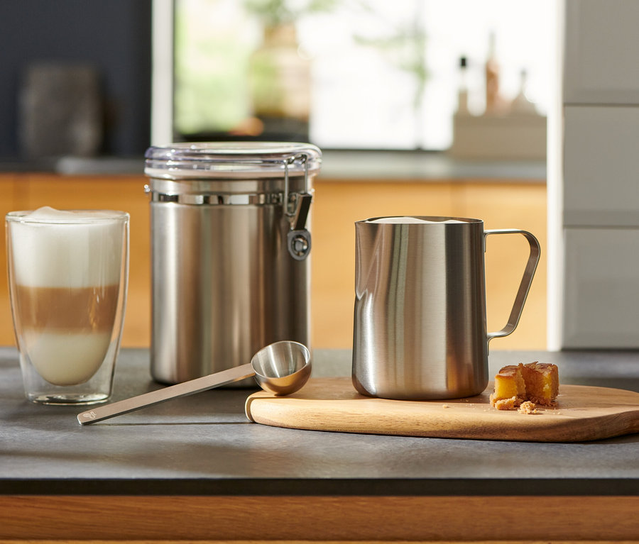 Auf der Küchentheke stehen ein Glas mit Latte Macchiato, ein silbernes Kaffeelot, ein Behälter und ein Milchkännchen.