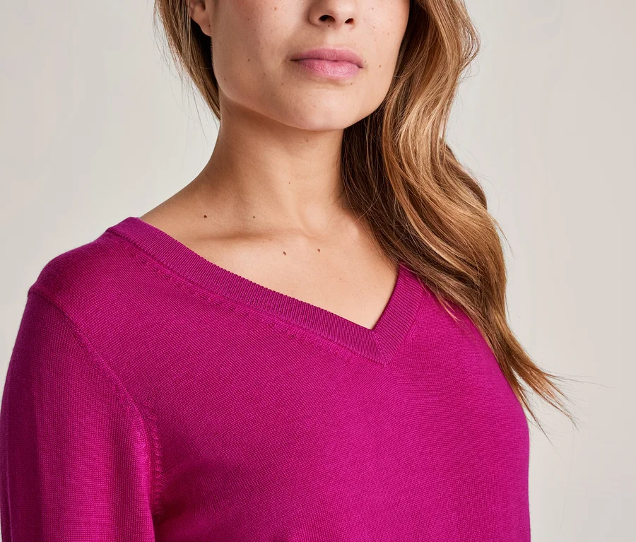Nahaufnahme einer Frau in einem fuchsiafarbenen V-Ausschnitt-Pullover, mit sichtbarem Kinn, Mund, Nase und langem, braunem, welligem Haar.