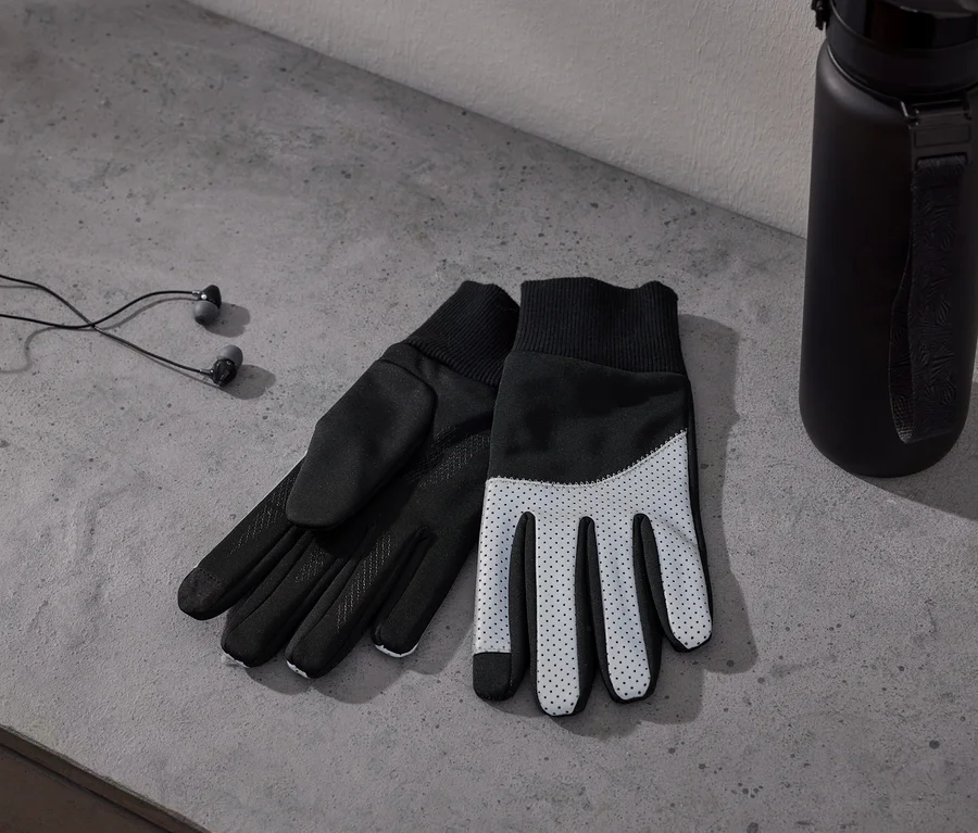 Zwei schwarz-weiße Handschuhe mit gepunkteten Handflächen, schwarze kabelgebundene Kopfhörer und eine schwarze Wasserflasche auf einer grauen Betonoberfläche.