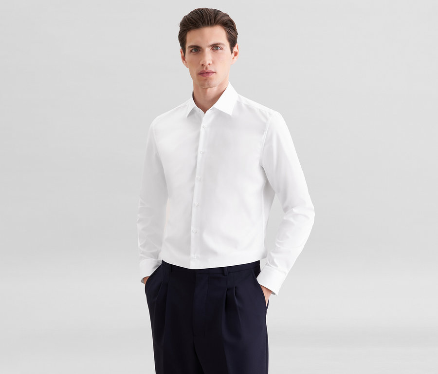 Mann trägt ein weißes Langarmhemd und eine dunkelblaue Bundfaltenhose, die Hände in den Hosentaschen.
