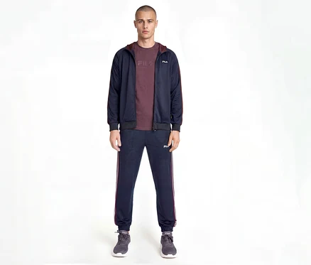 Mann trägt einen marineblauen Fila Trainingsanzug mit bordeauxroten Seitenstreifen, ein bordeauxrotes T-Shirt und graue Turnschuhe, stehend vor weißem Hintergrund.