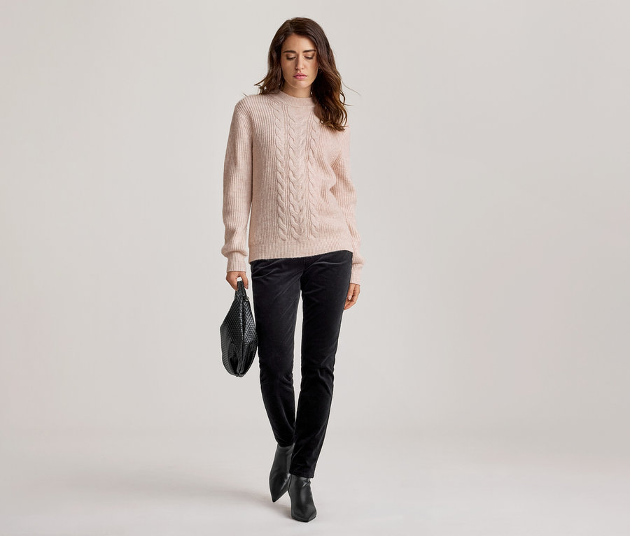 Frau in hellrosa Zopfstrickpullover, dunkler Samthose und schwarzen Stiefeletten, eine schwarze geflochtene Handtasche haltend, blickt nach unten.