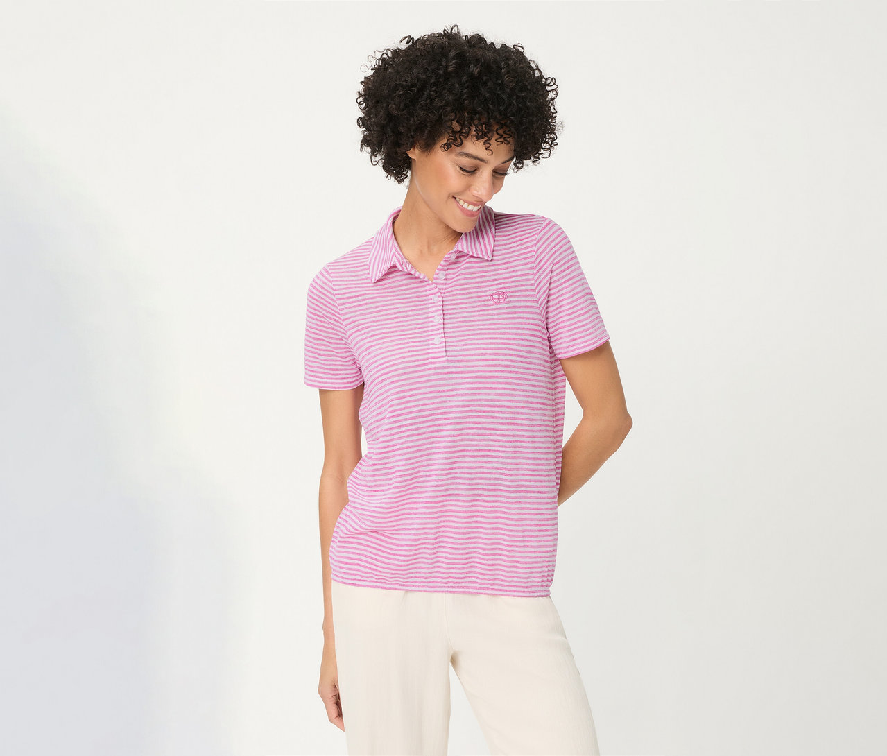 Lächelnde Frau mit lockigem Haar trägt ein pink gestreiftes Olsen Poloshirt und helle Hose, blickt nach unten.