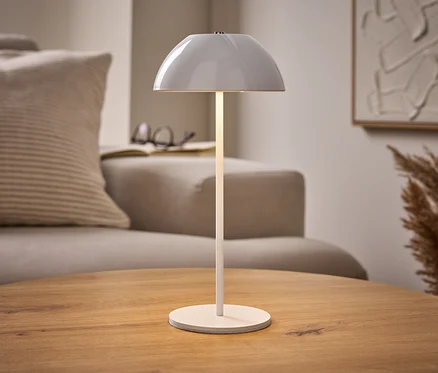 Beleuchtete weiße LED-Akku-Tischleuchte auf Holztisch, mit beigem Sofa, Buch und abstrakter Kunst im Hintergrund.
