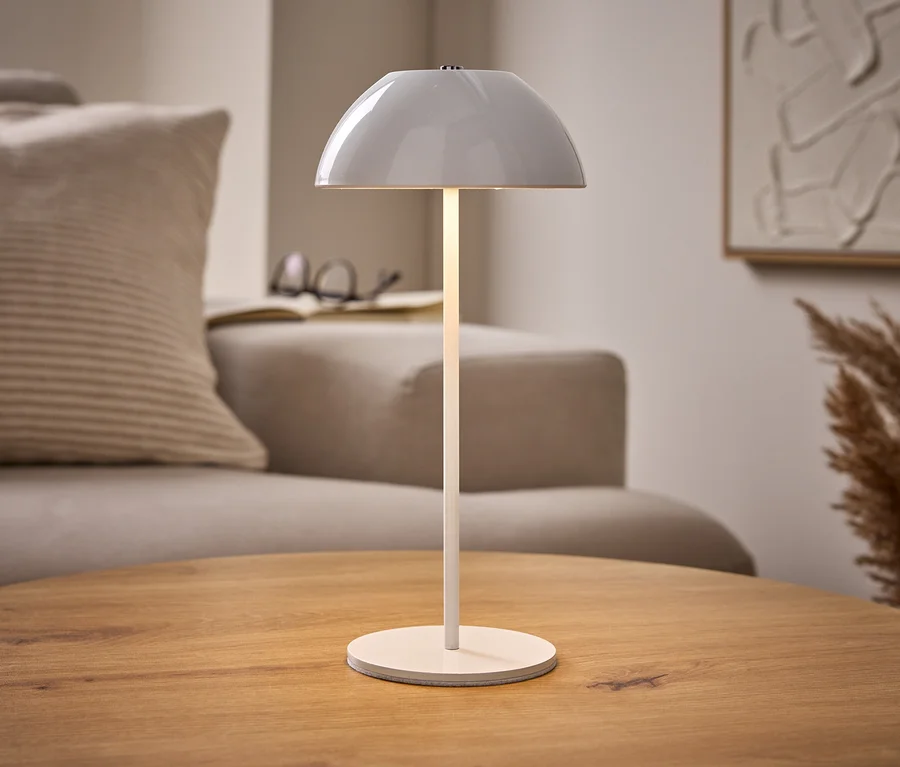 Beleuchtete weiße LED-Akku-Tischleuchte auf Holztisch, mit beigem Sofa, Buch und abstrakter Kunst im Hintergrund.