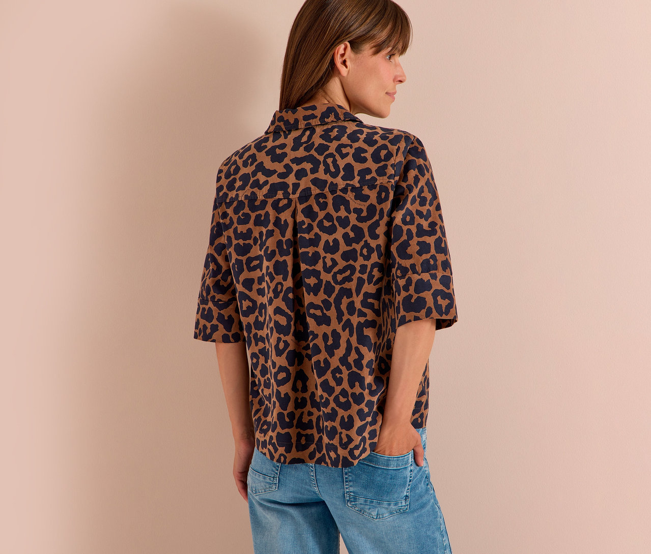 Frau von hinten, leicht gedreht, trägt eine braun-schwarze CECIL Hemdbluse mit Leopardenmuster und Jeans. Eine Hand steckt in der Hosentasche.