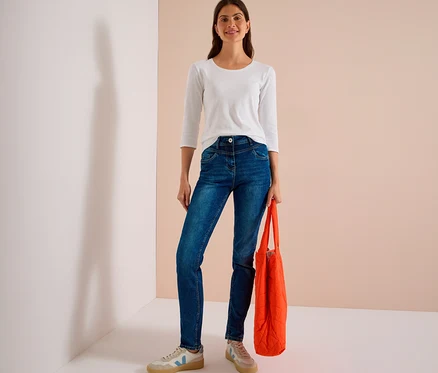 Lächelnde Frau trägt ein weißes Langarmshirt und blaue CECIL Jeans, hält eine orangefarbene Einkaufstasche.