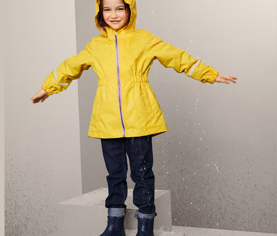 Ein Mädchen steht im Regen und trägt einen gelben Regenmantel aus recyceltem Material, Jeans und Gummistiefel.