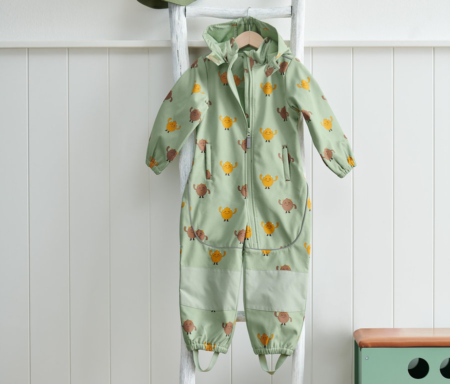 Grüner Softshell-Einteiler mit Monsterprint hängt an einer weißen Leiter neben einem grünen Hut.
