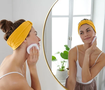 Frau reinigt Gesicht mit einem Wattepad und trägt zwei gelbe Kosmetik-Stirnbänder.