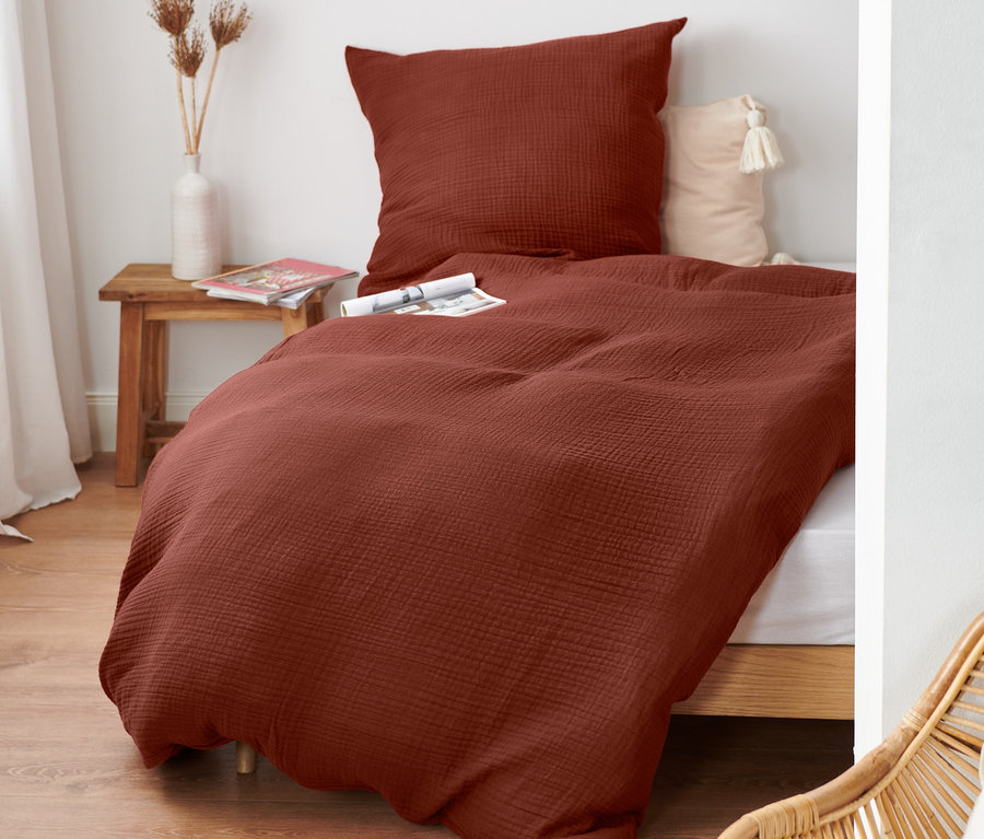 Rotbraune Musselin-Bettwäsche Normalgröße auf einem Bett mit Holzrahmen. Auf dem Nachttisch steht eine Vase mit Blumen.