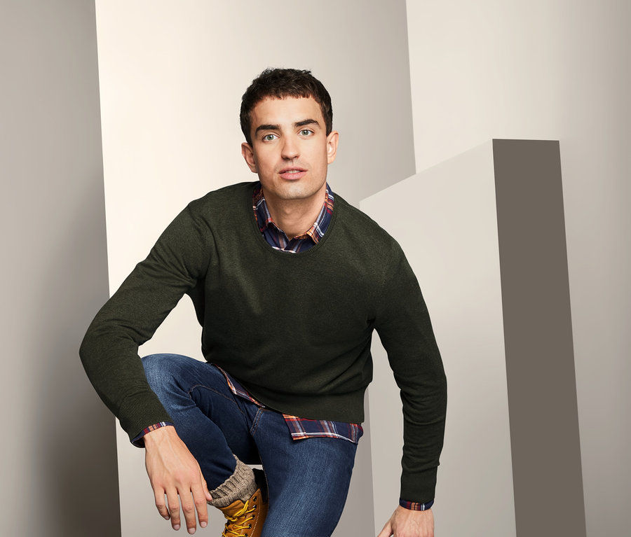 Mann posiert kniend und trägt einen Pullover mit Rundhalsausschnitt, Jeans und Stiefel.