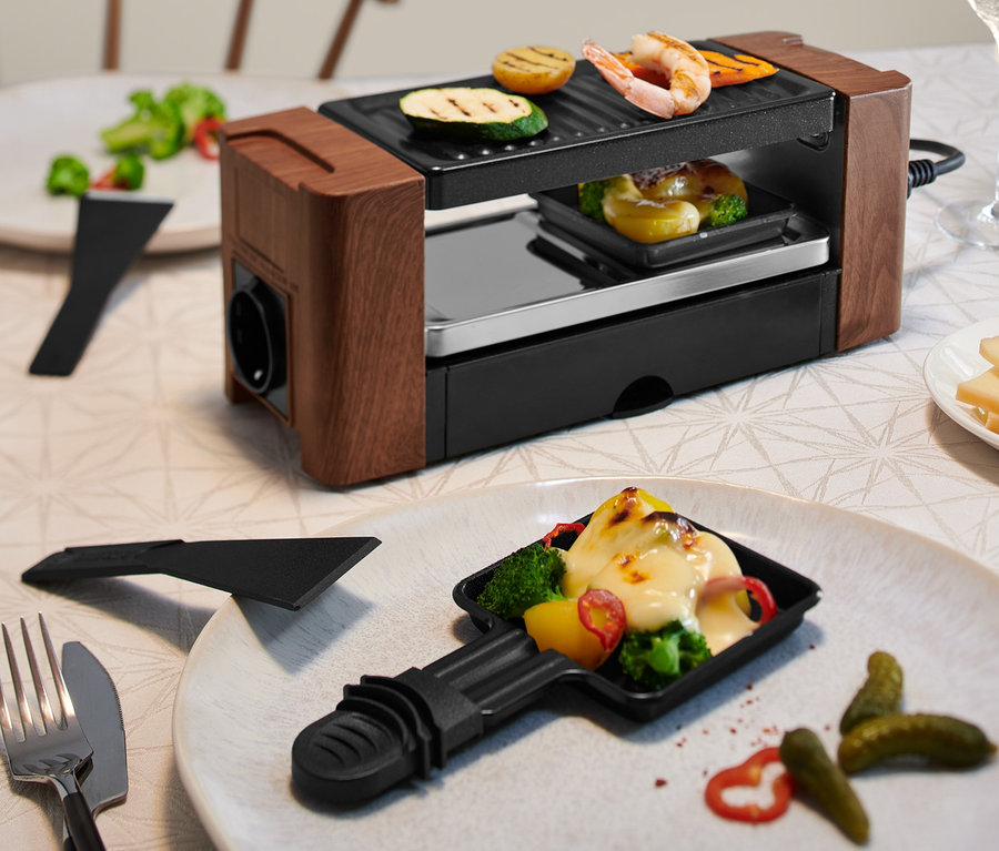 Auf einem Tisch steht ein Raclette für 2 Personen mit gegrilltem Gemüse und Käse.