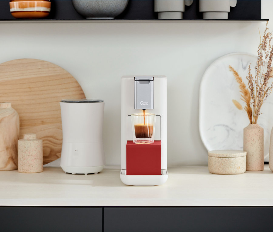 Auf einer Küchentheke steht eine Qbo Kaffeemaschine, die Kaffee in ein Glas brüht. Zu sehen sind eine Qbo ICONIC Dark Chrome und eine Qbo ESSENTIAL Bright Coral.