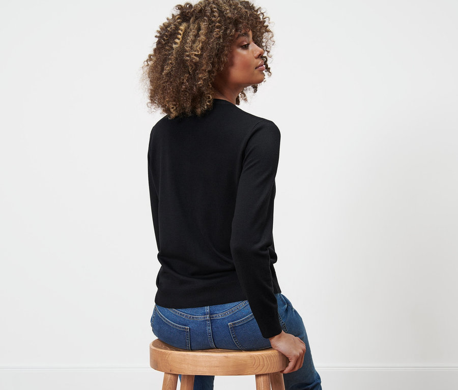 Frau sitzt seitlich auf einem Holzhocker, trägt einen Feinstrick-Cardigan und Jeans.