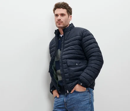 Mann lehnt an Wand, trägt marineblaue Steppjacke und Jeans.