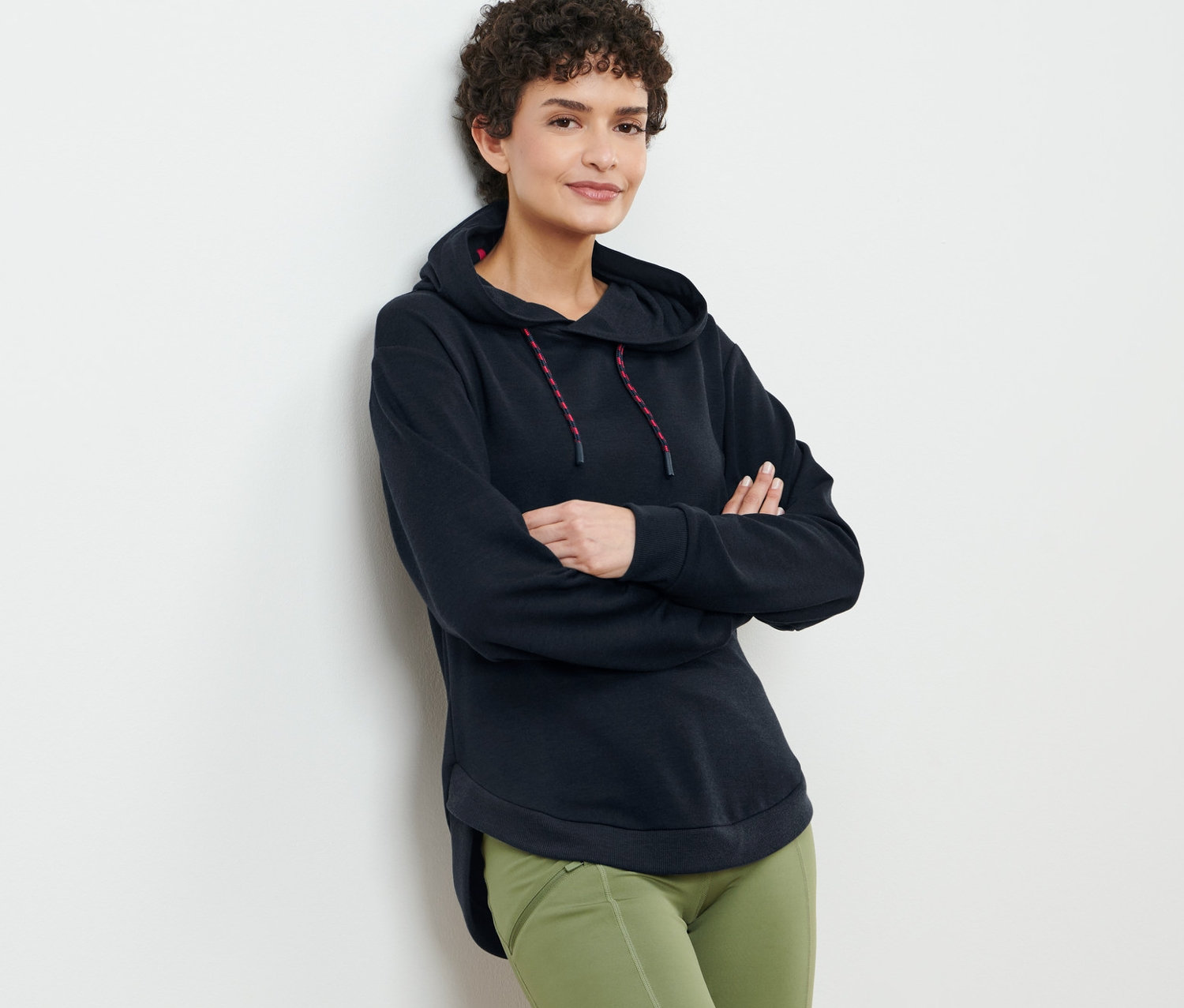 Frau mit braunem lockigem Haar posiert in schwarzem Kapuzen-Sweatpullover und grüner Hose.