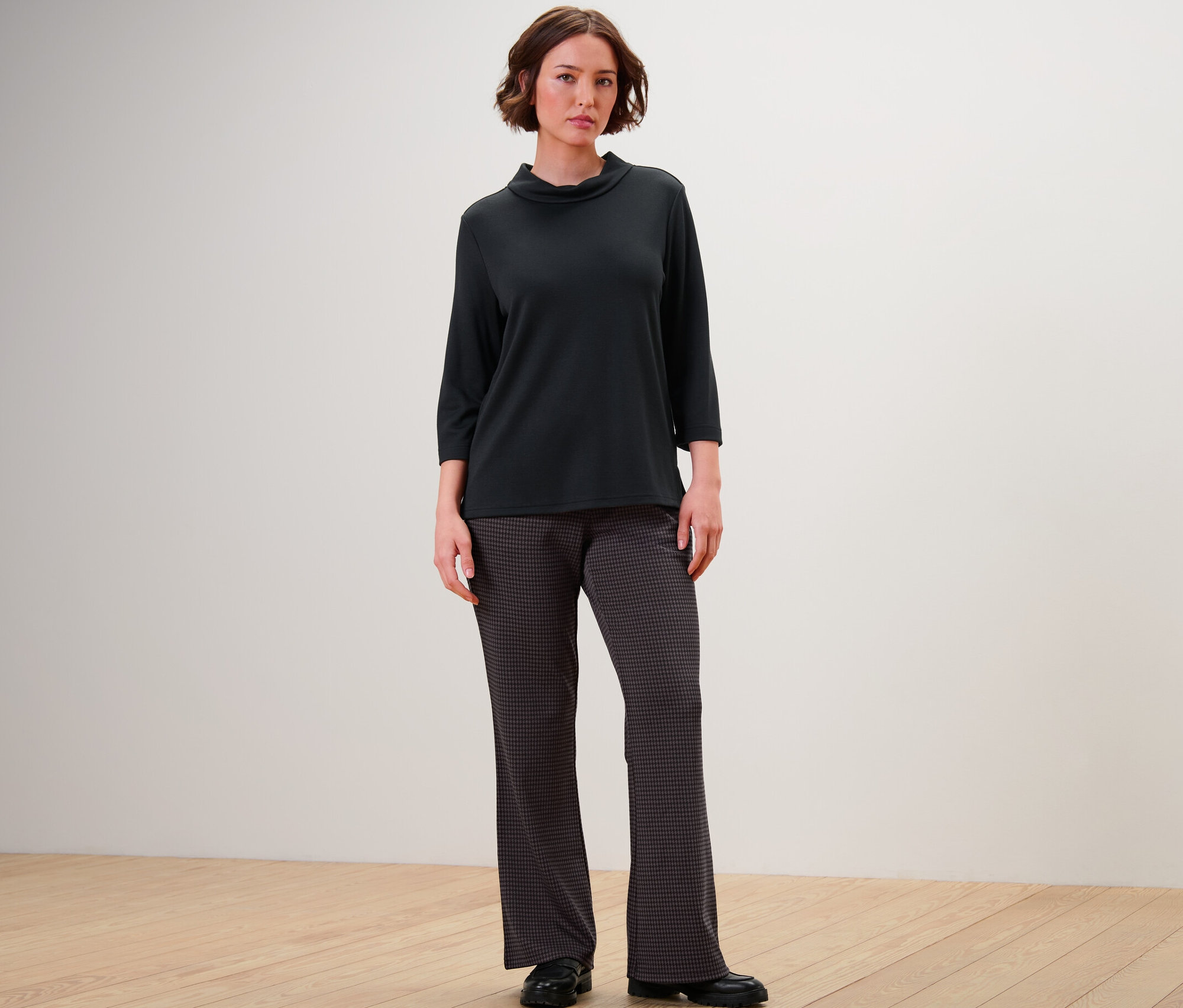 Eine Frau steht und trägt ein schwarzes Shirt mit Dreiviertelärmeln und eine Schlupfhose mit Hahnentritt-Jacquard.