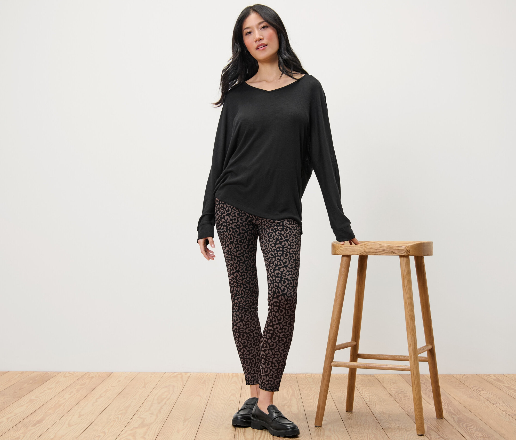 Frau steht neben einem Holzhocker und trägt ein schwarzes Langarmshirt und bedruckte Heavy-Leggings.