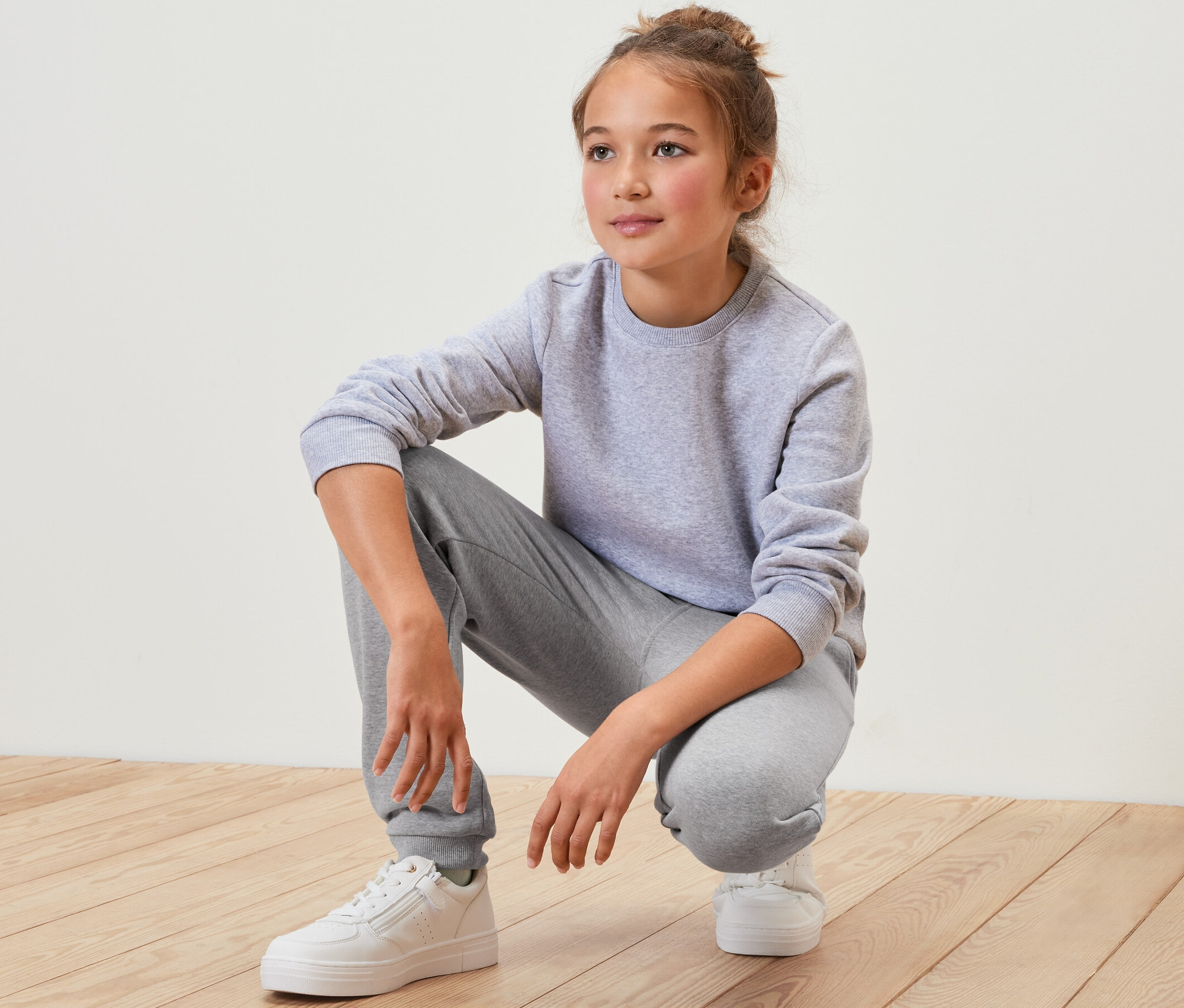 Ein Mädchen in einem grauen Trainingsanzug und weißen Schuhen hockt auf einem Holzboden. Sie trägt eine Kinder-Sweathose, grau.