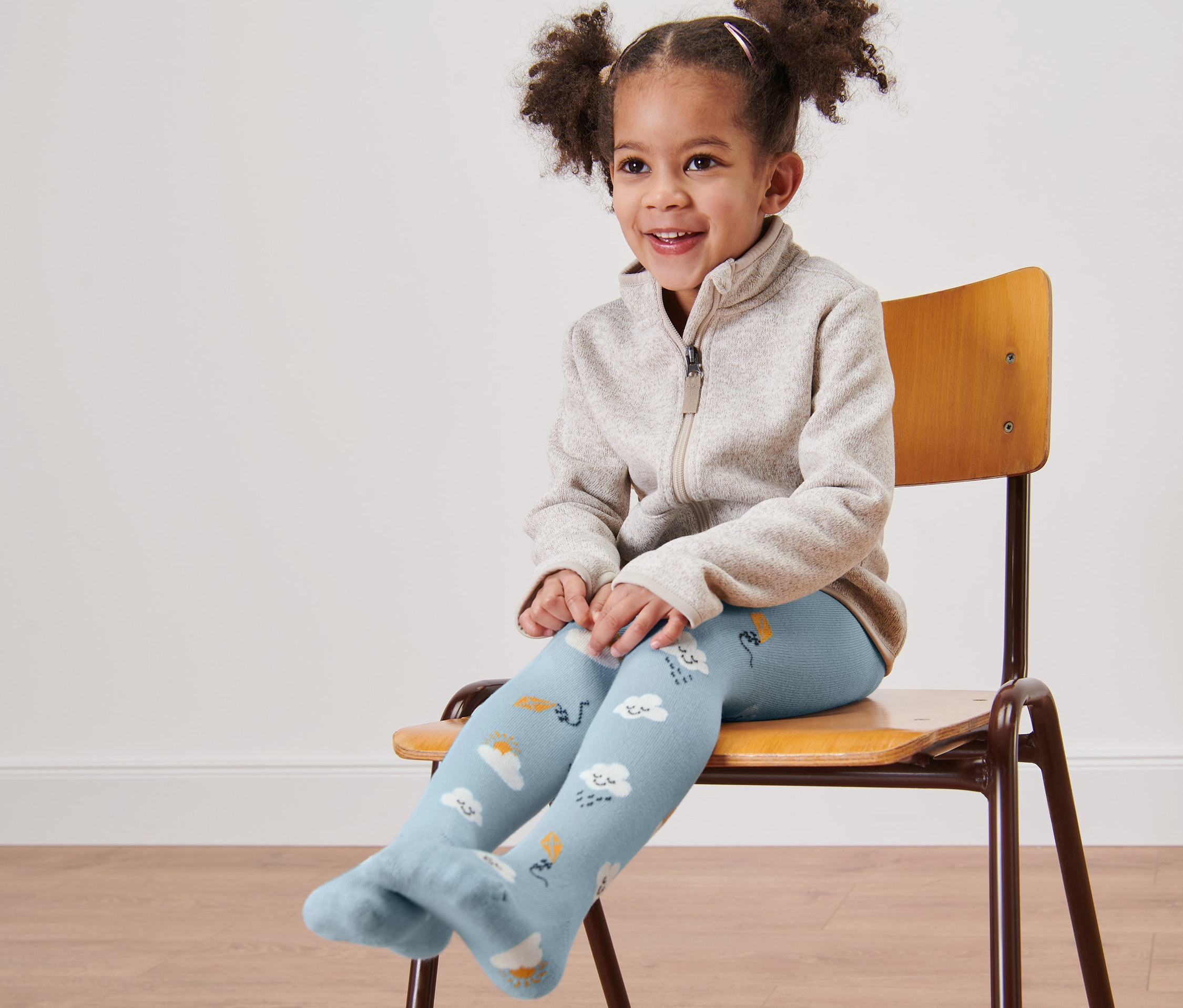 Lächelndes kleines Mädchen sitzt auf einem Stuhl und trägt blaue Strumpfhosen mit Wolken.