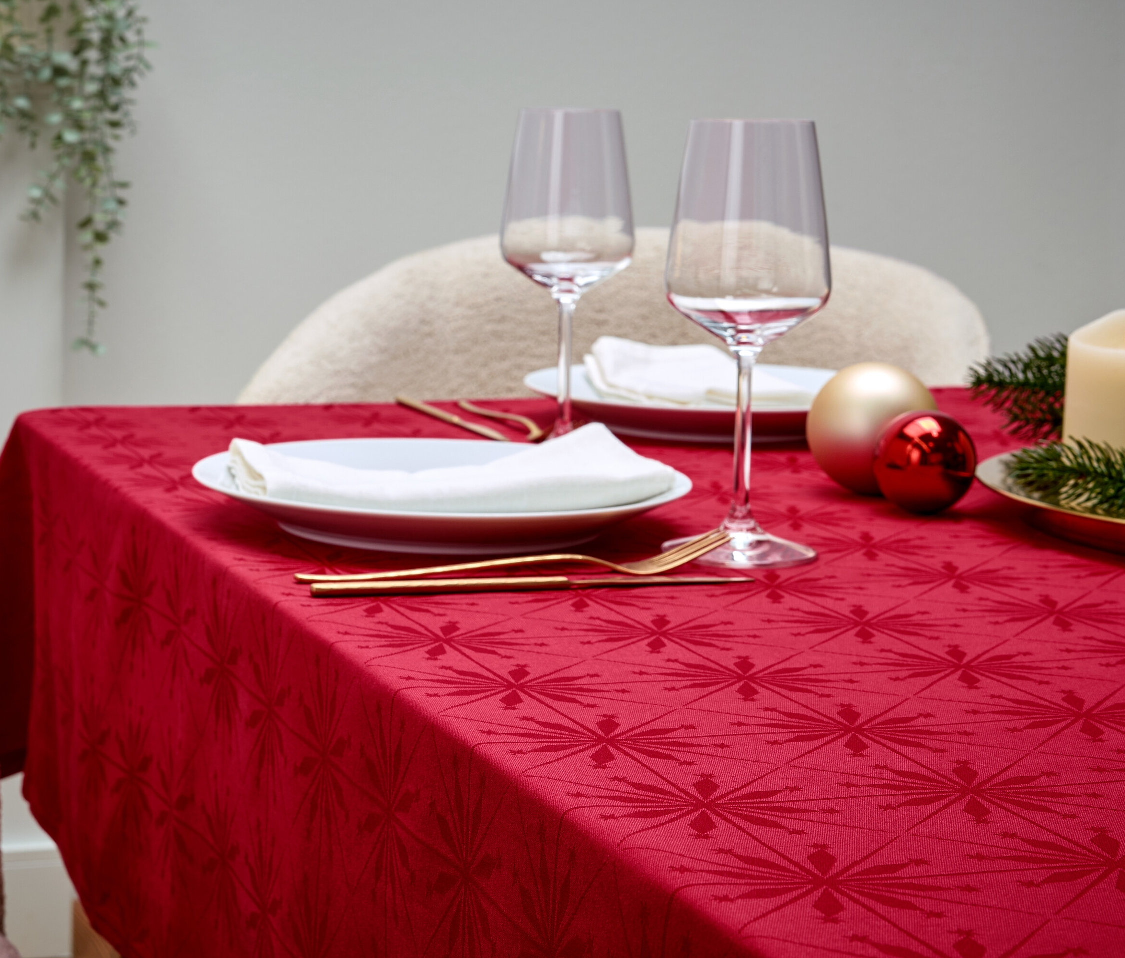 Ein gedeckter Tisch mit roter Tischdecke, weißen Tellern, Weingläsern, goldenem Besteck, Dekorationen und einer Kerze.