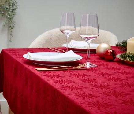 Ein gedeckter Tisch mit roter Tischdecke, weißen Tellern, Weingläsern, goldenem Besteck, Dekorationen und einer Kerze.