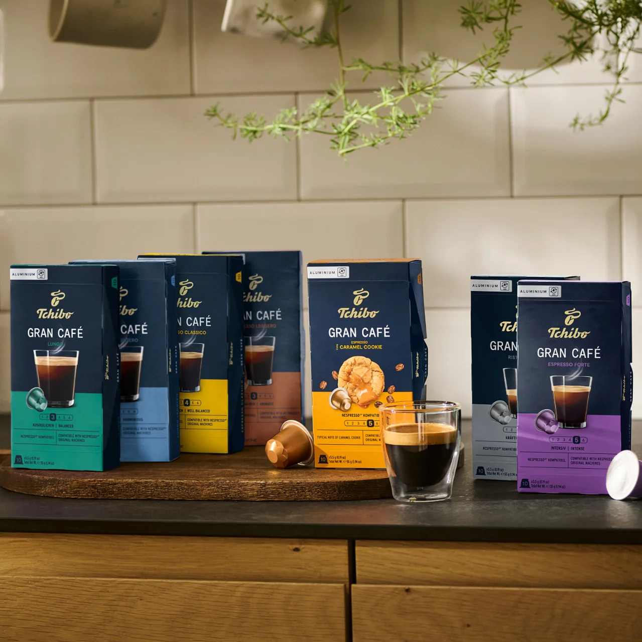 Auf einem Holztablett stehen Packungen Tchibo Gran Café Kaffeekapseln verschiedener Geschmacksrichtungen und ein Glas Kaffee.