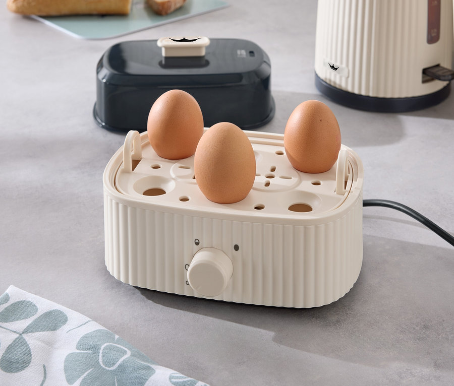 Drei Eier in einem Eierkocher auf einer Küchentheke, mit einem Toaster, einem Wasserkocher und Brot im Hintergrund.