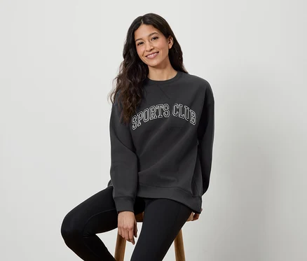 Frau sitzt auf einem Hocker, trägt ein dunkelgraues Sweatshirt mit der Aufschrift SPORTS CLUB und schwarze Leggings.