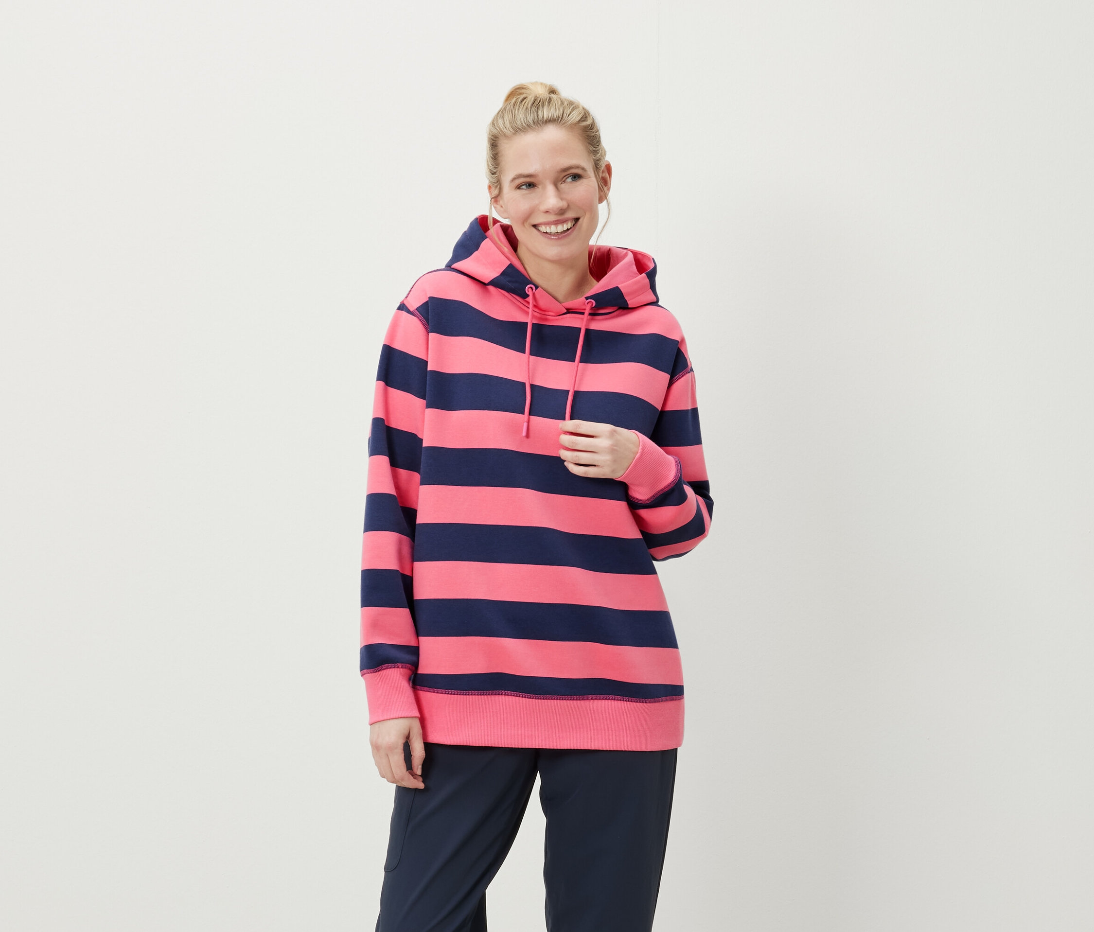 Lächelnde Frau mit blonden Haaren trägt einen pink-blau gestreiften Hoodie und dunkelblaue Hosen.