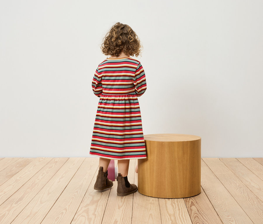 Mädchen in gestreiftem Kleid steht mit dem Rücken zu einem zylindrischen Tischchen und trägt braune Stiefel.