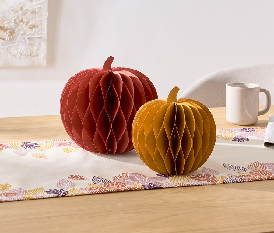 Auf einem Tisch stehen zwei dekorative Papierkürbisse, ein roter und ein gelber. Daneben befinden sich eine Tasse und eine Serviette.