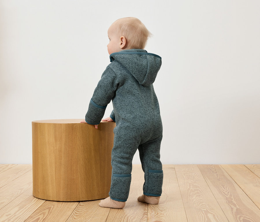 Ein Baby in einem blauen Overall stützt sich auf einen runden Holztisch.