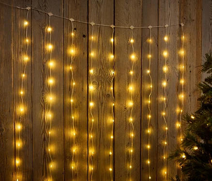 Aufgehängte Lichterketten vor einem Holzhintergrund neben einem Weihnachtsbaum.