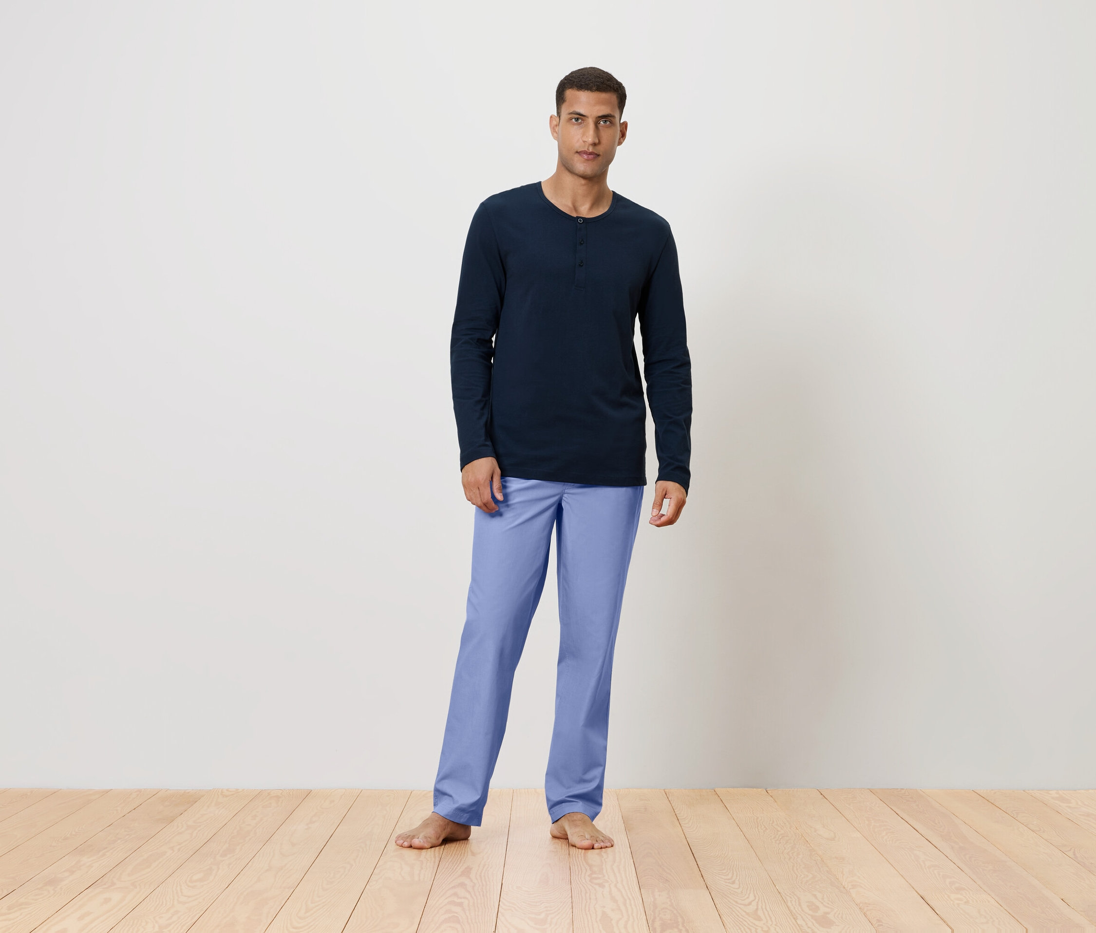 Mann steht barfuß auf einem Holzboden und trägt ein dunkelblaues Langarmshirt und hellblaue Hosen.
