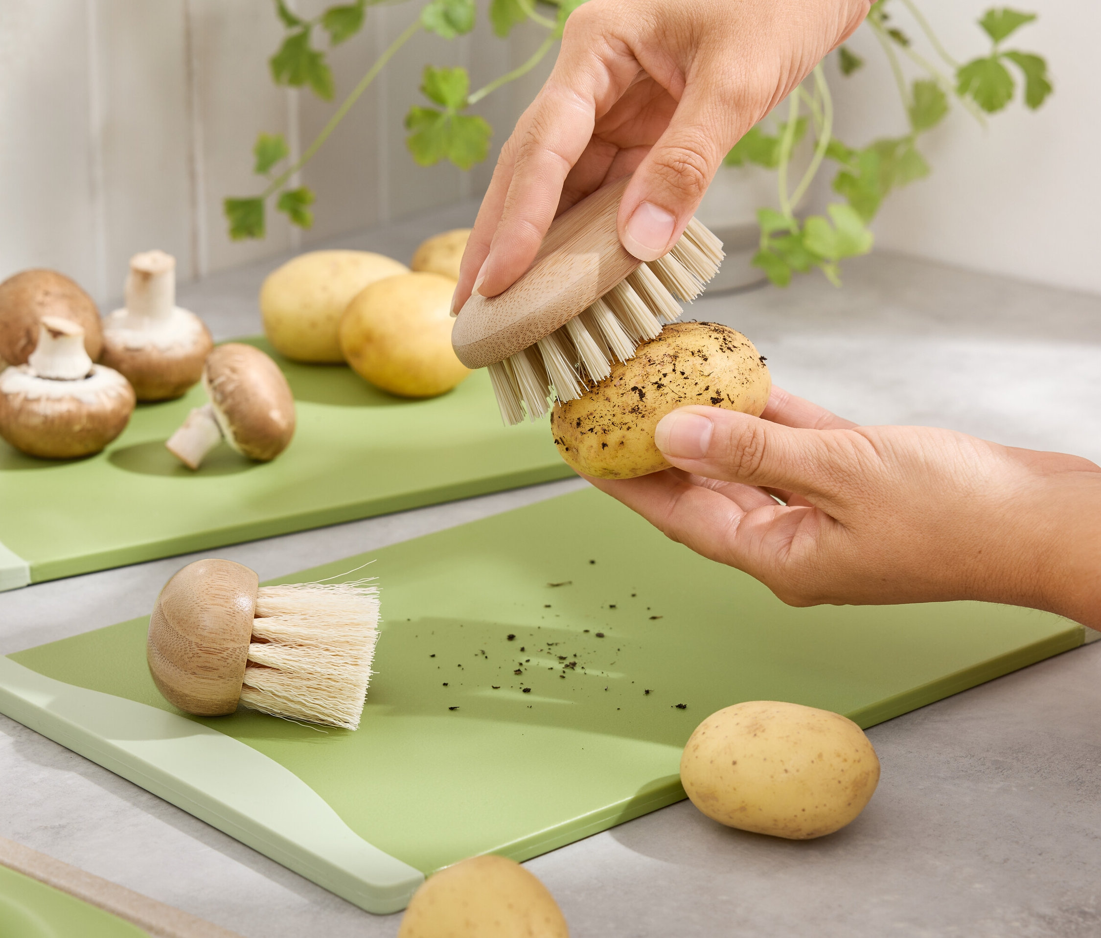 Hände putzen eine Kartoffel mit einer Bürste auf einem grünen Schneidebrett. Braune Champignons und Kartoffeln sind im Hintergrund.