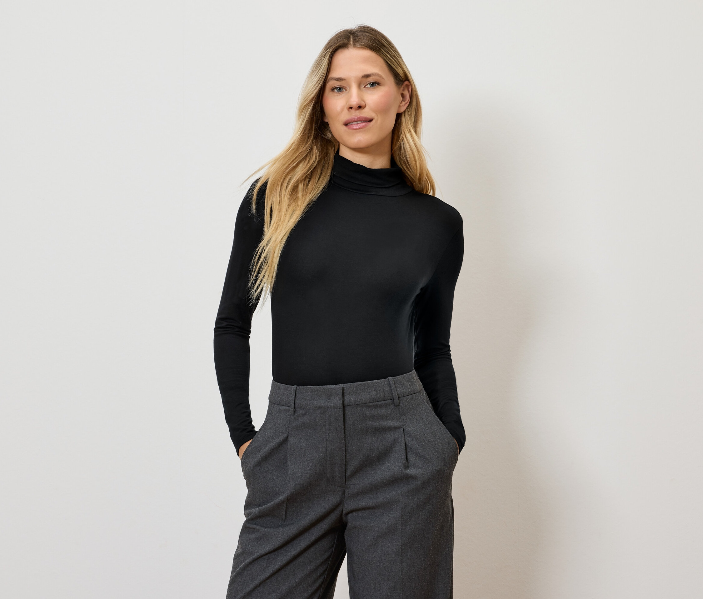 Frau mit langen blonden Haaren posiert in einem schwarzen Rollkragenpullover und einer grauen Hose.