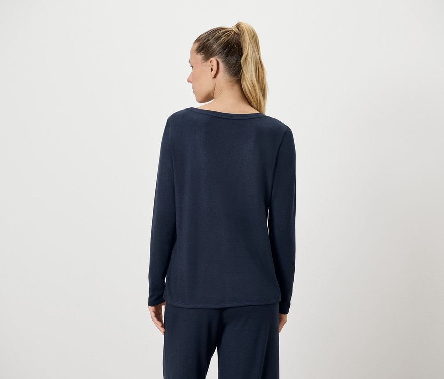 Von hinten ist eine Frau mit blonden Haaren zu sehen, die zu einem Pferdeschwanz zusammengebunden sind. Sie trägt ein dunkelblaues Langarmshirt und eine Hose.