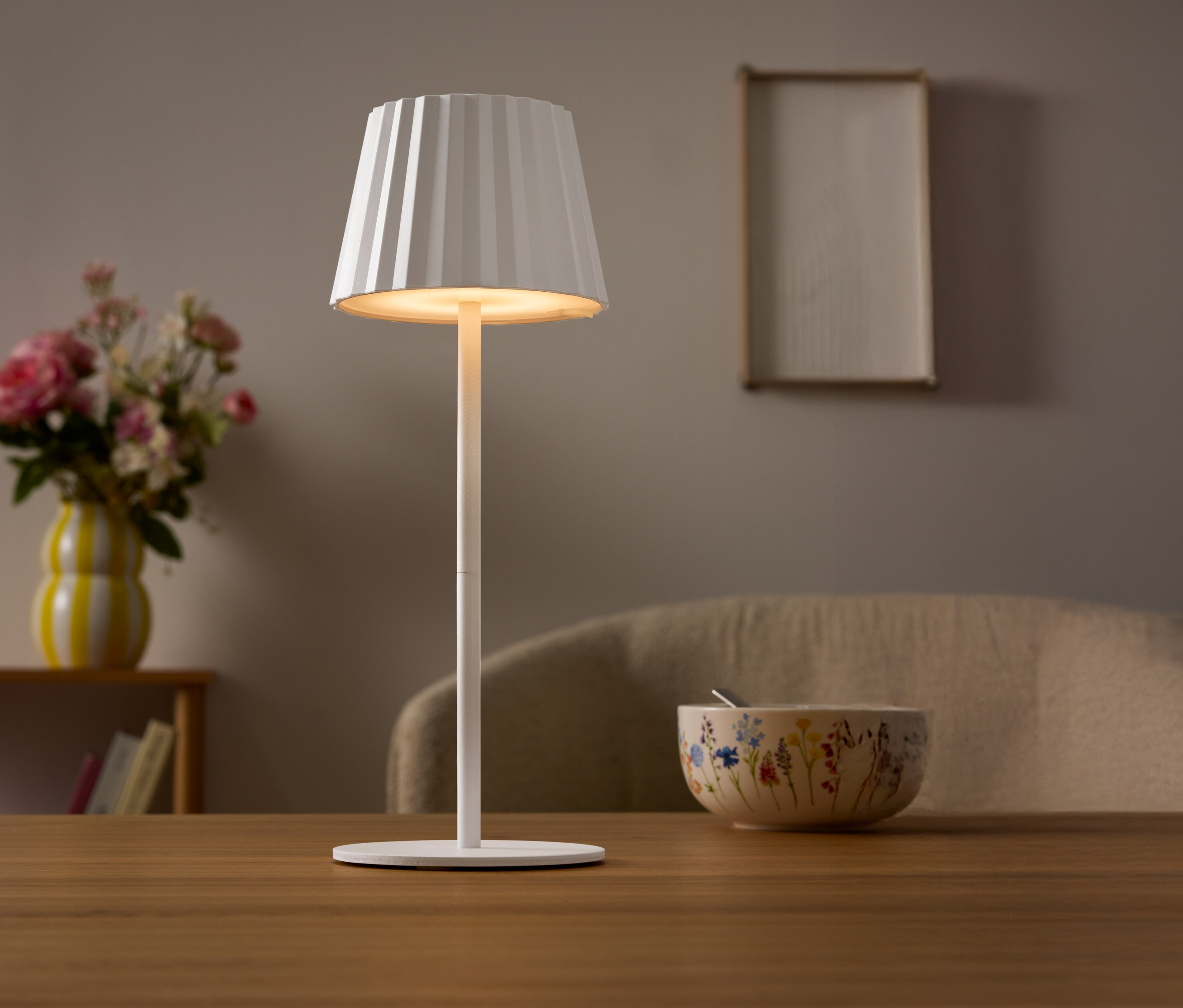 Auf einem Tisch steht eine weiße Lampe mit einem plissierten Lampenschirm. Im Hintergrund befinden sich eine Blumenvase und eine Schüssel.