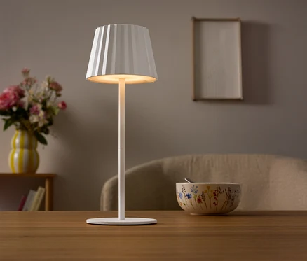 Auf einem Tisch steht eine weiße Lampe mit einem plissierten Lampenschirm. Im Hintergrund befinden sich eine Blumenvase und eine Schüssel.